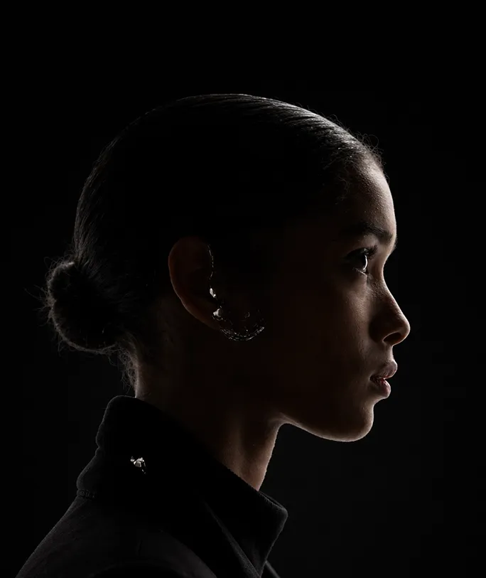 Seitenprofil eines jungen Menschen mit zurückgebundenem Haar und dunklem Oberteil vor schwarzem Hintergrund.