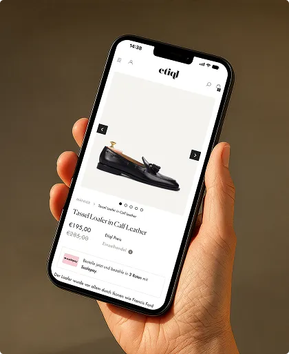 Hand hält ein Smartphone, das eine Produktseite für schwarze Tassel-Loafer aus Kalbsleder auf der Website etiql zeigt.