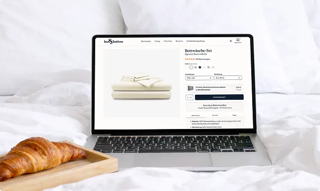 Laptop auf weißem Bett, zeigt eine Website mit einem beige Bettwäsche-Set und einem aufgeklappten Croissant auf einem Holztablett daneben.