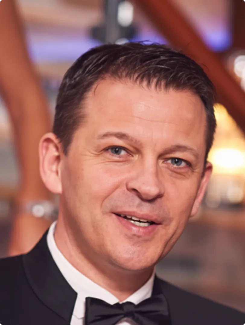 Lächelnder Mann im schwarzen Smoking mit Fliege vor unscharfem Hintergrund.