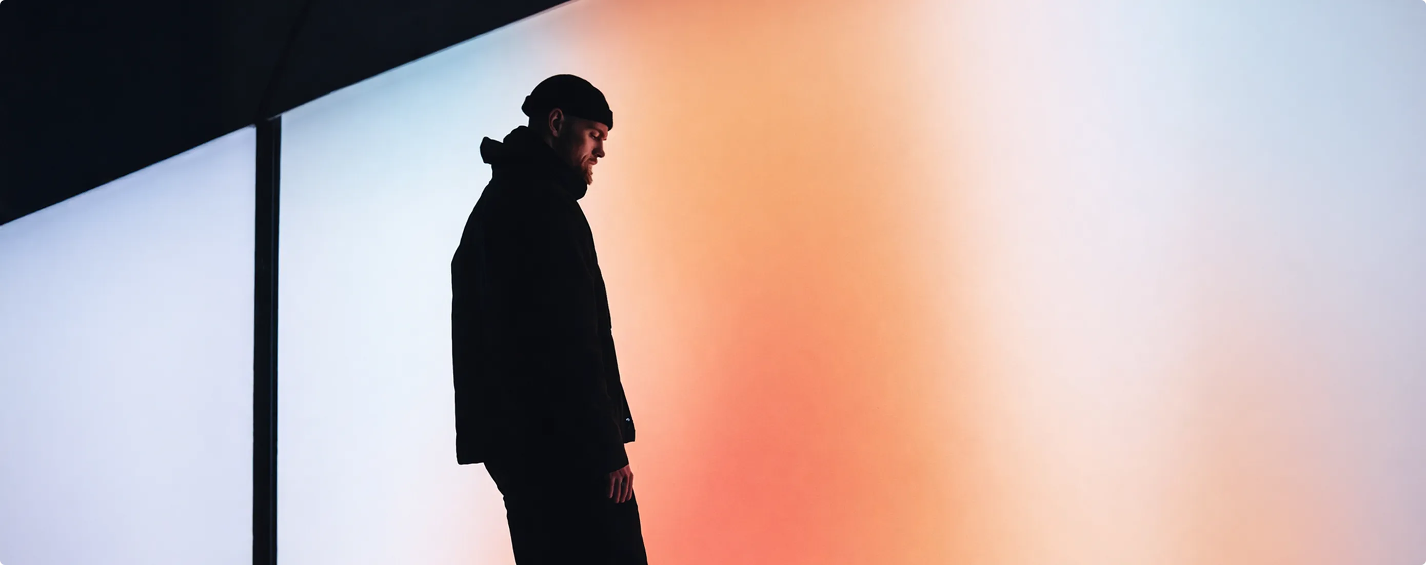 Mann in schwarzer Kleidung steht vor einer Wand mit orange-weißem Farbverlauf.