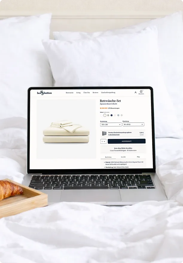 Laptop auf einem Bett mit weißen Kissen zeigt eine Website für ein ausverkauftes Bettwäsche-Set in Cremefarbe.