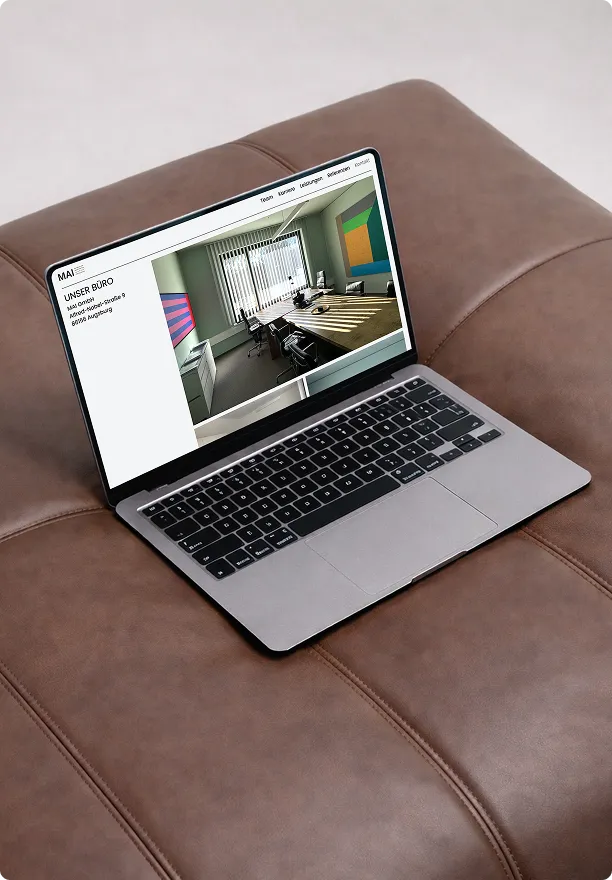 Ein geöffneter Laptop liegt auf einem braunen Ledersofa, auf dem Bildschirm ist eine Büro-Webseite mit einem Bild eines modernen Konferenzraums zu sehen.