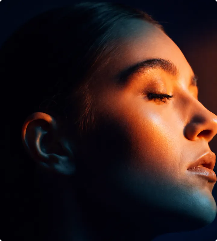 Seitliches Porträt einer entspannten Frau mit geschlossenen Augen, beleuchtet von warmem Licht vor dunklem Hintergrund.