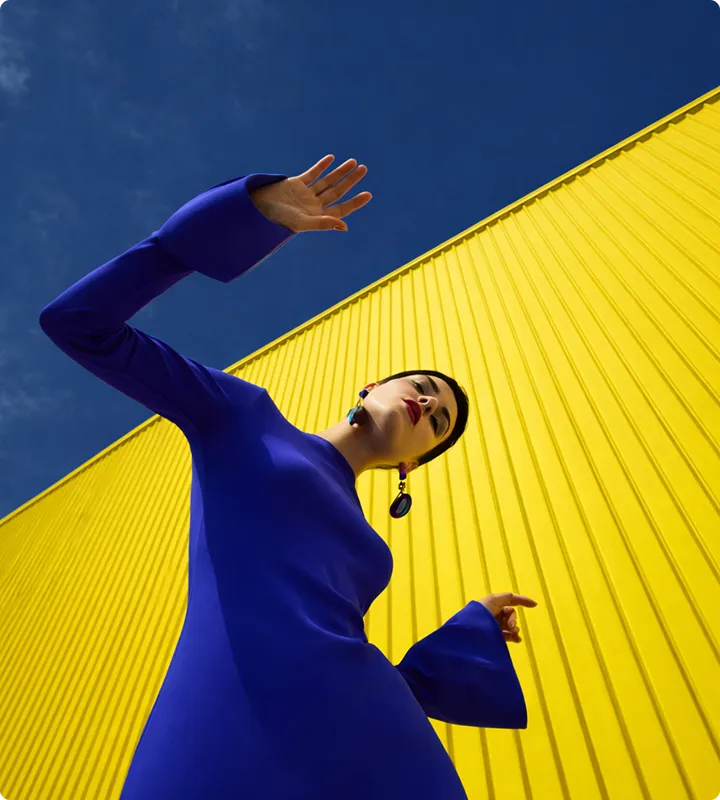 Frau in blauem Kleid posiert vor einer gelben Wand vor blauem Himmel.