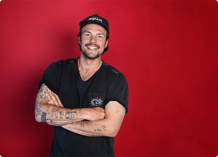 Junger Mann mit schwarzen T-Shirt und Baseballkappe, mit verschränkten Armen vor rotem Hintergrund lächelnd.