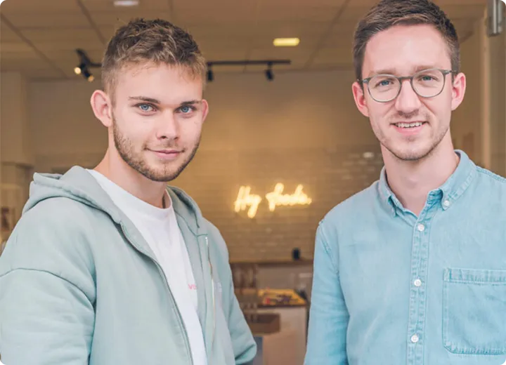Zwei junge Männer, einer mit Kapuzenjacke und Bart, der andere mit Brille und hellblauem Hemd, lächeln in die Kamera vor einem unscharfen Hintergrund mit Schriftzug.