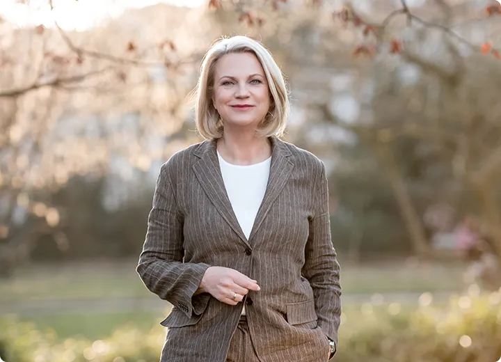 Lächelnde blonde Frau in gestreiftem Blazer und weißem Shirt steht draußen in einem Park mit weichem, verschwommenem Hintergrund.