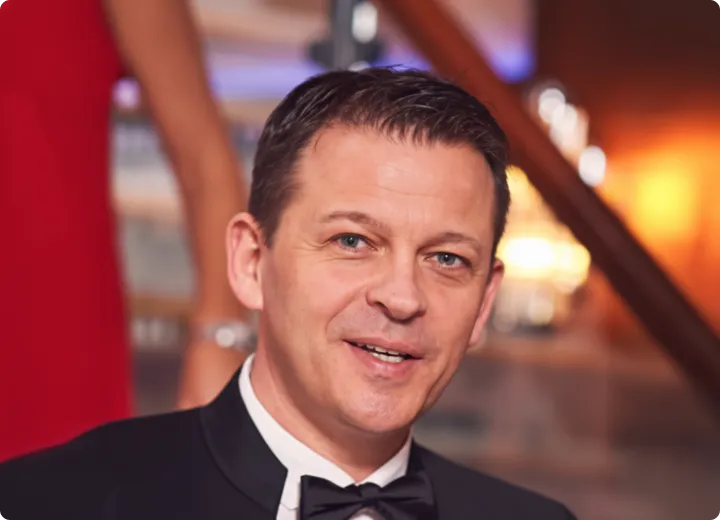 Porträt eines lächelnden Mannes in schwarzer Smokingjacke mit Fliege bei einer festlichen Veranstaltung.