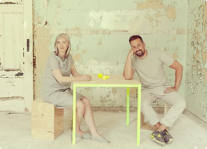 Paar sitzt an einem einfachen Holztisch in einem Raum mit abgeblätterten, hellgrünen Wänden.