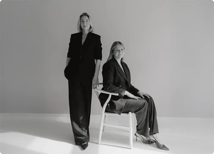 Schwarz-weiß-Foto von zwei Frauen in eleganter Anzugmode, eine steht mit Händen in den Taschen, die andere sitzt auf einem Holzstuhl.