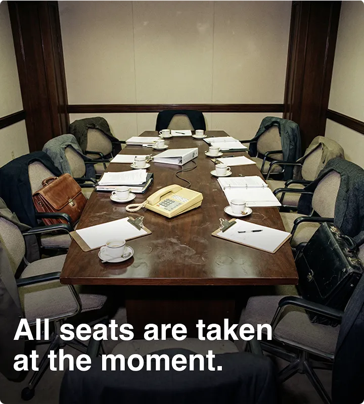 Ein Besprechungsraum mit einem großen Holztisch, auf dem Kaffeetassen, Aktenordner, Notizblöcke und ein Telefon stehen, umgeben von Stühlen mit Mänteln und Aktentaschen, die darauf hinweisen, dass alle Plätze besetzt sind.