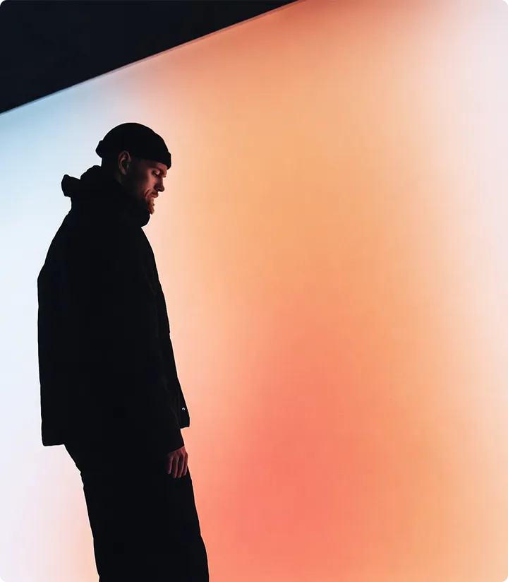 Ein Mann mit schwarzer Kleidung und Mütze steht seitlich vor einem Hintergrund mit einem Farbverlauf von hellblau zu orange.