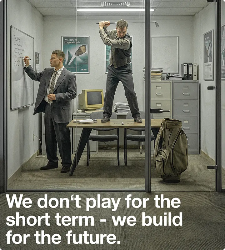 Zwei Männer in einem Büro; einer schreibt an ein Whiteboard, während der andere auf einem Tisch steht und Golfschwung ausführt, neben einem Golfbag.