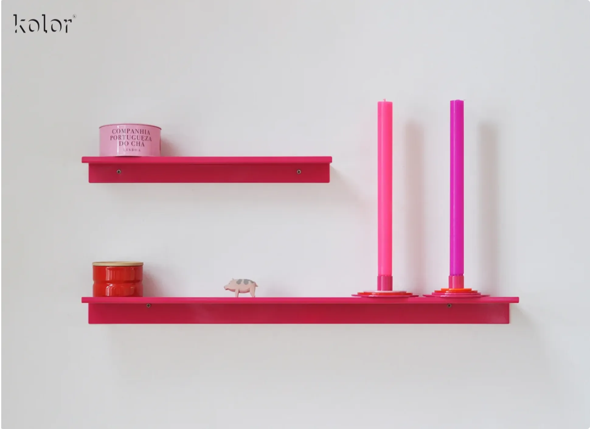 Zwei pinke Regalbretter an einer weißen Wand mit einer rosa Dose, einer roten Dose, einer kleinen Schweinfigur und zwei hohen pinkfarbenen Kerzen.