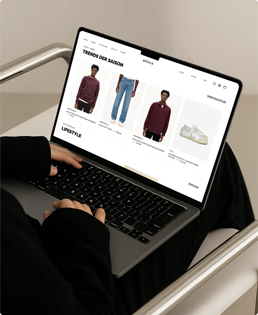 Person mit schwarzer Jacke benutzt Laptop mit einer Mode-Webseite, die saisonale Trends wie Pullover, Jeans, Hemd und weiße Sneaker zeigt.