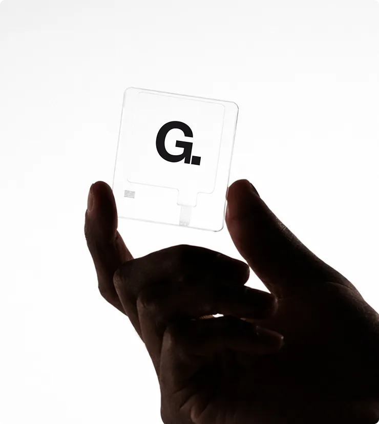 Dunkle Hand hält transparentes Quadrat mit dem Buchstaben ‚G.‘ vor hellem Hintergrund.