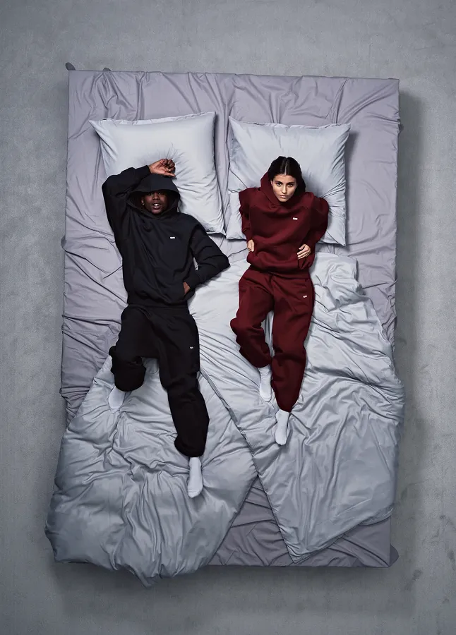 Zwei Personen liegen auf einem Bett mit grauer Bettwäsche; eine trägt schwarze, die andere dunkelrote Freizeitkleidung.