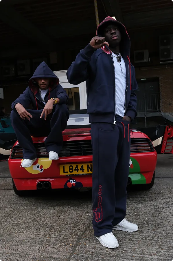 Zwei Männer mit Kapuzenjacken und Jogginghosen posieren vor einem roten Sportwagen mit gelben und grünen Aufklebern in einer Garage.