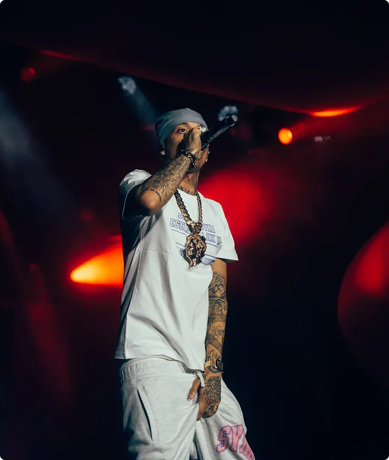 Mann mit Tattoos und grauer Mütze trägt weißen T-Shirt und Jogginghose, singt mit Mikrofon auf roter Bühnenbeleuchtung.