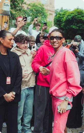 Ein Mann in einem pinken Outfit und Sonnenbrille lächelt und schüttelt jemandem die Hand, während eine Gruppe von Menschen mit Handys ihn fotografiert.
