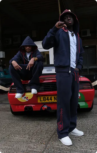 Zwei Männer in dunklen Hoodies und Freizeitkleidung posieren vor einem roten Auto mit bunten Kindermotiven.