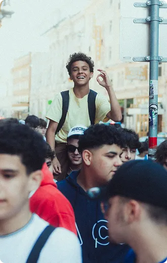 Junger Mann mit Rucksack, der auf den Schultern einer anderen Person sitzt und lächelt, umgeben von einer Gruppe junger Leute in einer Stadt.