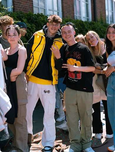 Gruppe junger Menschen posiert draußen vor einem Gebäude, zwei junge Männer in der Mitte zeigen Handzeichen und lächeln in die Kamera.