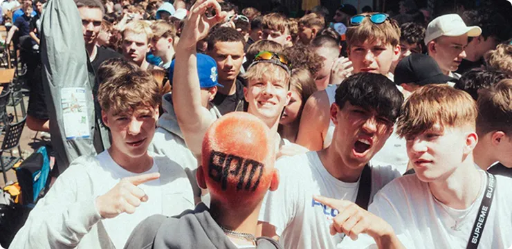 Junge Gruppe in Menschenmenge, drei von ihnen zeigen auf einen Mann mit orange gefärbtem Haar und dem Schriftzug 'epm' am Hinterkopf.