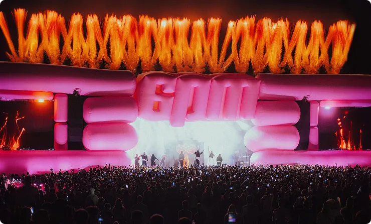 Große Menschenmenge bei einem Konzert vor einer aufblasbaren Bühne mit pinken Händen und Feuerwerk im Hintergrund.