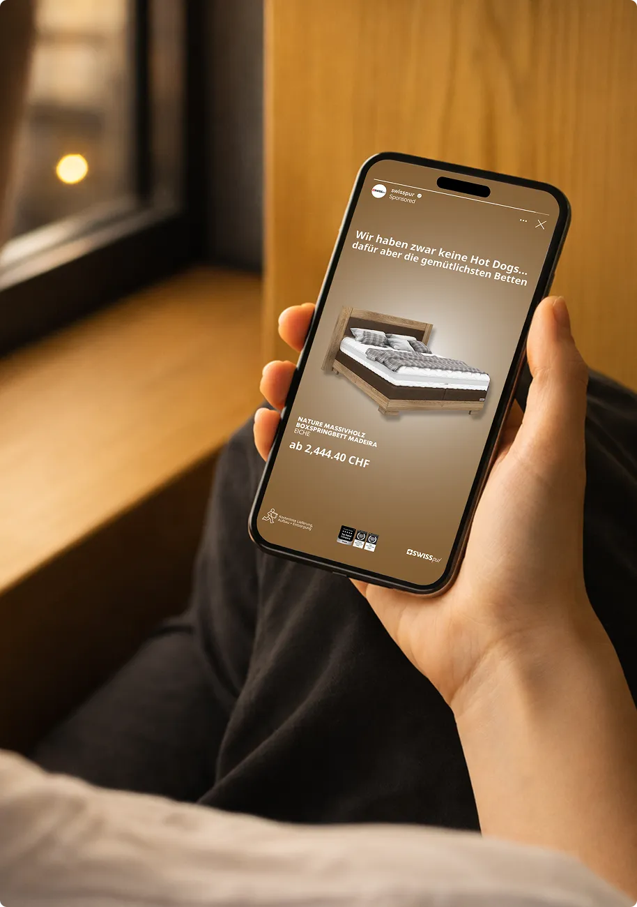 Hand hält ein Smartphone mit einer Anzeige für ein massives Holzbett namens Madeira, Preis ab 2.444,40 CHF.