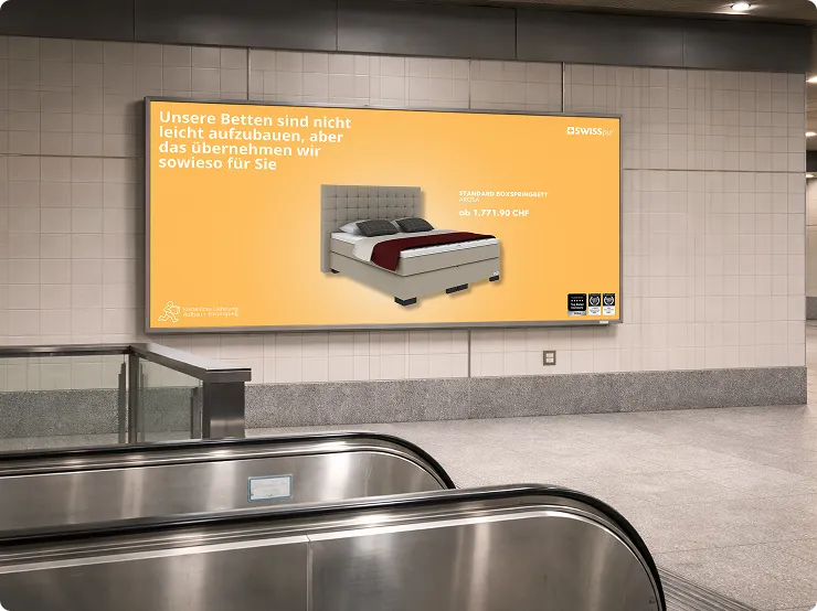 Werbetafel in einer U-Bahn-Haltestelle mit einem Bild eines Bettes und dem Text: Unsere Betten sind nicht leicht aufzubauen, aber das übernehmen wir sowieso für Sie. Preis: ab 1.771,90 CHF.