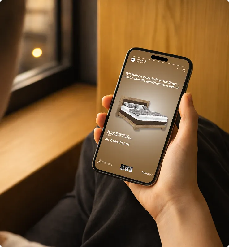 Person hält ein Smartphone, auf dem eine Anzeige für ein Bett aus Massivholz mit dem Text „Wir haben zwar keine Hot Dogs, dafür aber die gemütlichsten Betten“ zu sehen ist.