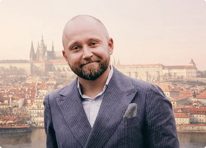 Porträt eines lächelnden Mannes mit Bart und Anzug vor einer Stadtlandschaft mit historischen Gebäuden und einem Fluss.