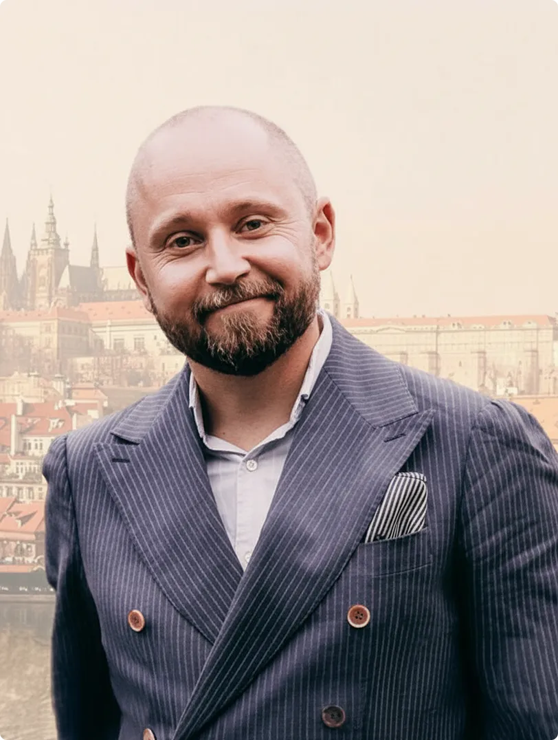 Mann mit Glatze und Bart trägt einen gestreiften Anzug mit weißem Hemd und einem gestreiften Einstecktuch vor einer historischen Stadtkulisse.