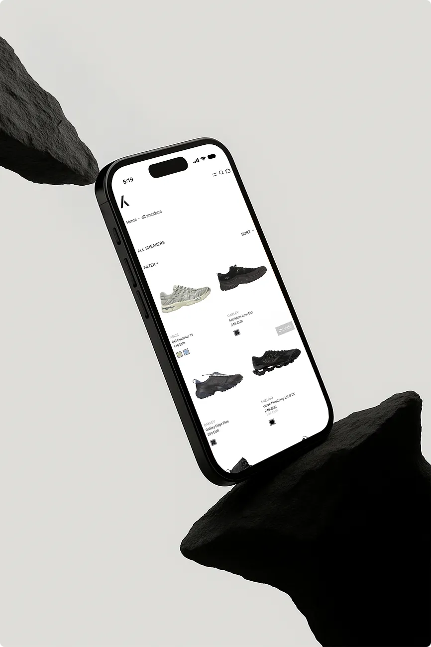 Smartphone zeigt eine Online-Shop-Seite mit vier verschiedenen Sneakermodellen auf weißem Hintergrund zwischen schwarzen Felsen.