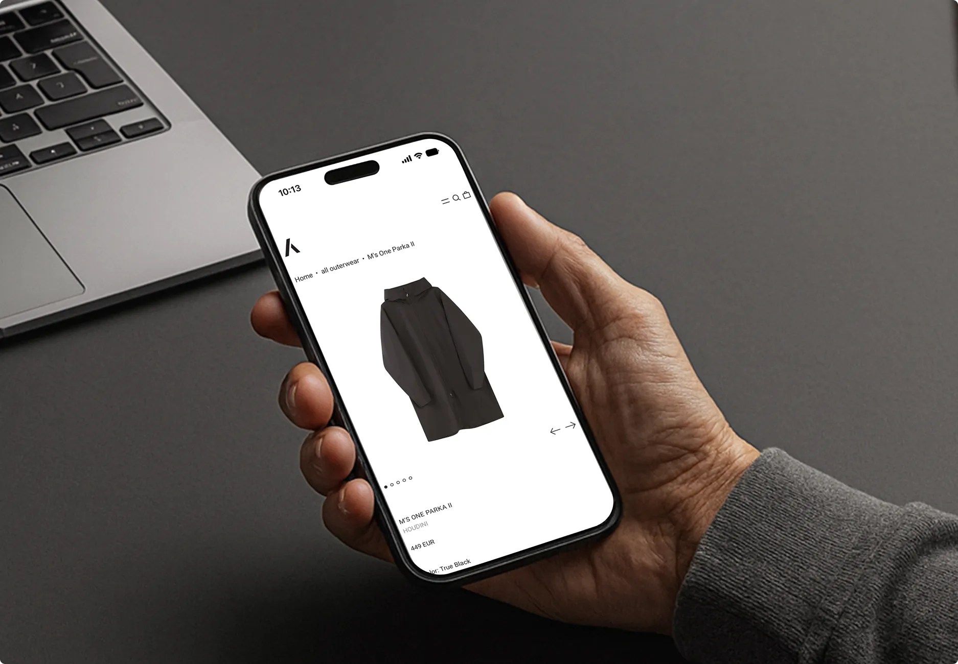Hand hält ein Smartphone, das eine Produktseite für eine schwarze M'S One Parka II Jacke zeigt, daneben liegt ein Laptop.