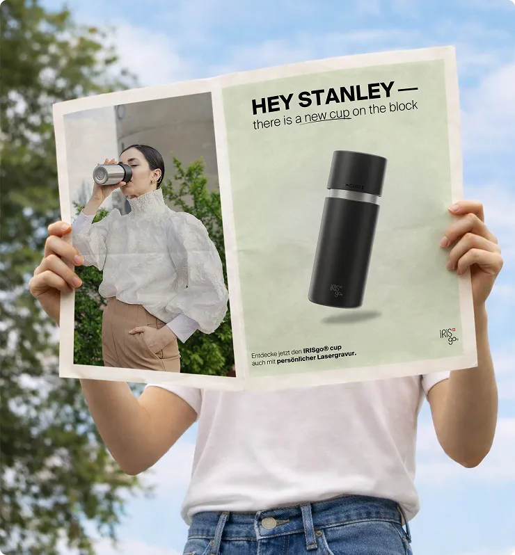 Person hält eine Zeitung mit einer Anzeige für einen schwarzen IRISgo® Trinkbecher und einem Foto einer Frau, die aus einem ähnlichen Becher trinkt.