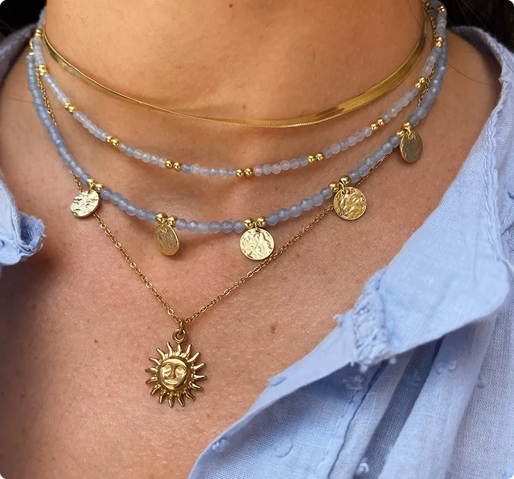 Close-up of a neck with several gold and light blue pearl necklaces, as well as a sun pendant necklace and a light blue shirt.