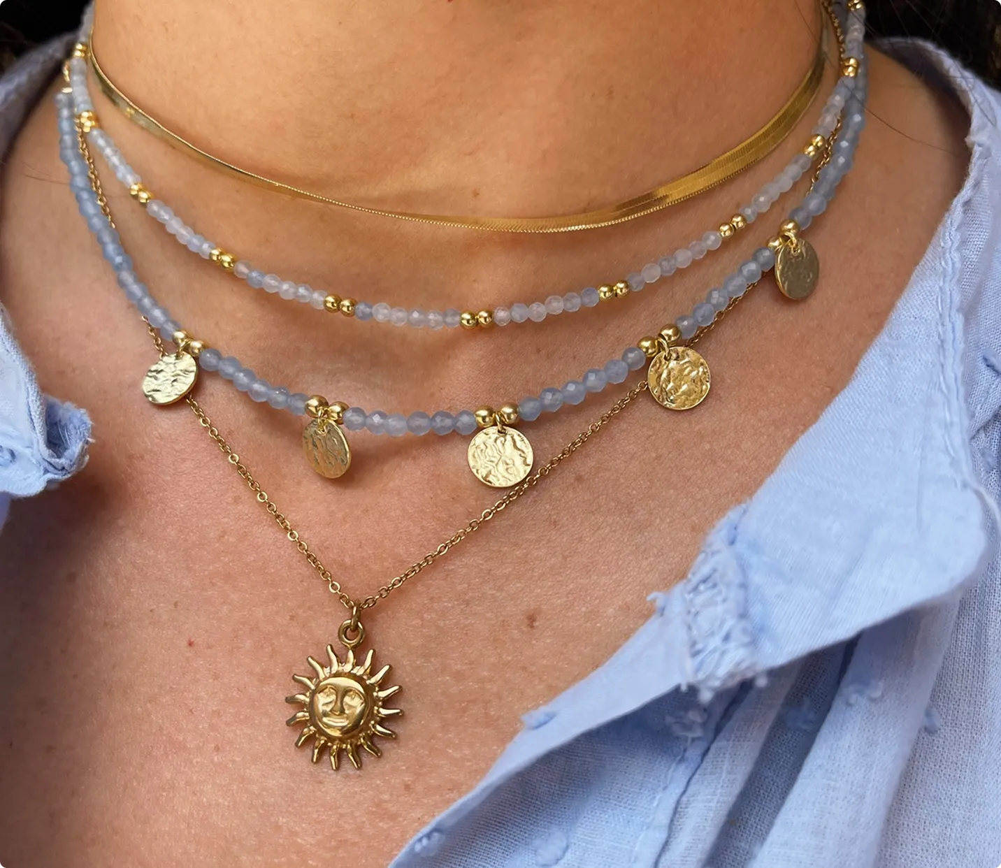 Multi-layered necklace with gold coin charms and a gold sun pendant on a person wearing a light blue shirt.