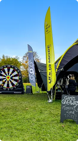 Outdoor event setup with an inflatable dartboard, two vertical banners, a tent, and a sign offering games, giveaways, and tomorrow's land tickets.