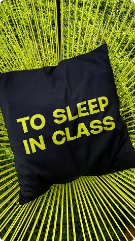 Schwarzes Kissen mit gelber Aufschrift 'TO SLEEP IN CLASS' auf einem gelben Stuhl mit Streben.