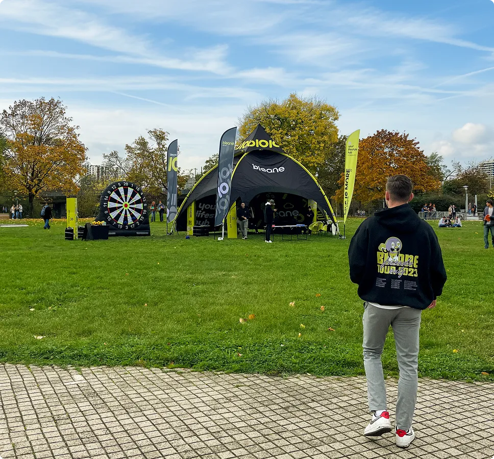 Junger Mann mit 'Bleaone Tour 2023' Kapuzenpullover auf einem gepflasterten Weg vor einem Parkstand mit Werbezelt, Luftdartscheibe und Bannern in herbstlicher Landschaft.