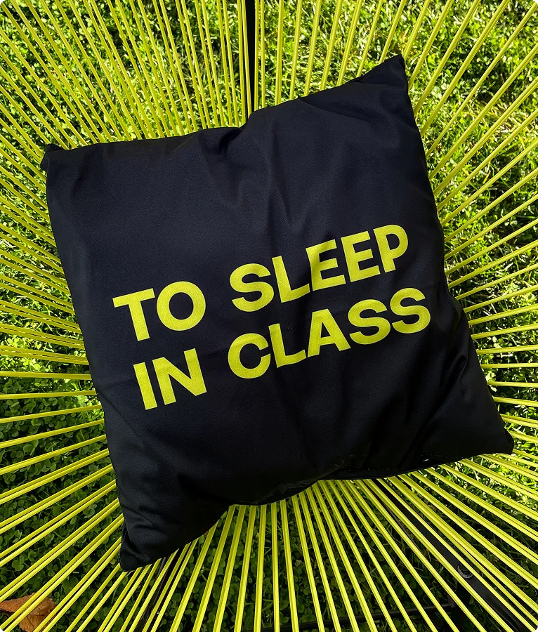 Schwarzes Kissen mit gelber Aufschrift ‚TO SLEEP IN CLASS‘ auf einem gelben Stuhl im Freien.