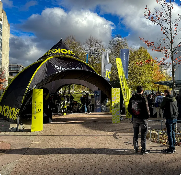 Outdoor event with a black and yellow tent and banners, people gathered around on a paved area under a partly cloudy sky.