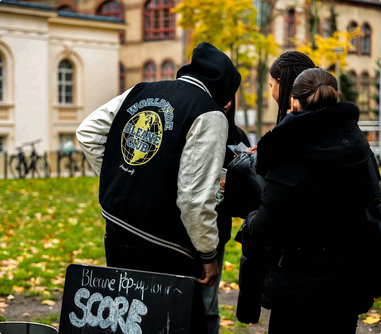 Person mit schwarzer und weißer Jacke mit Aufschrift ‚Worldwide Bleane Club‘ auf dem Rücken spricht mit zwei anderen Personen im Freien vor einer Tafel mit der Aufschrift ‚Bleane Pop-up Tour SCORE‘.