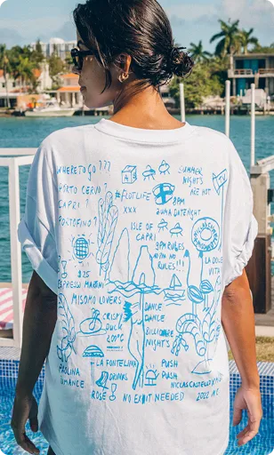 Frau mit dunklen Haaren im Dutt trägt weißes T-Shirt mit blauen, handschriftlichen Reisezeichnungen und Texten auf dem Rücken, steht vor Wasser und Häusern.