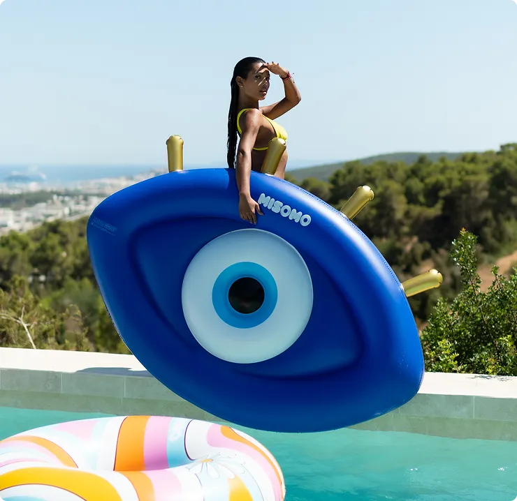 Frau im gelben Bikini hält ein großes, blaues aufblasbares Schwimmspielzeug in Form eines Auges am Pool.