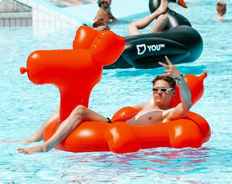 Junger Mann liegt entspannt auf einem großen orangefarbenen aufblasbaren Entenfloß in einem Schwimmbad und zeigt ein Peace-Zeichen.