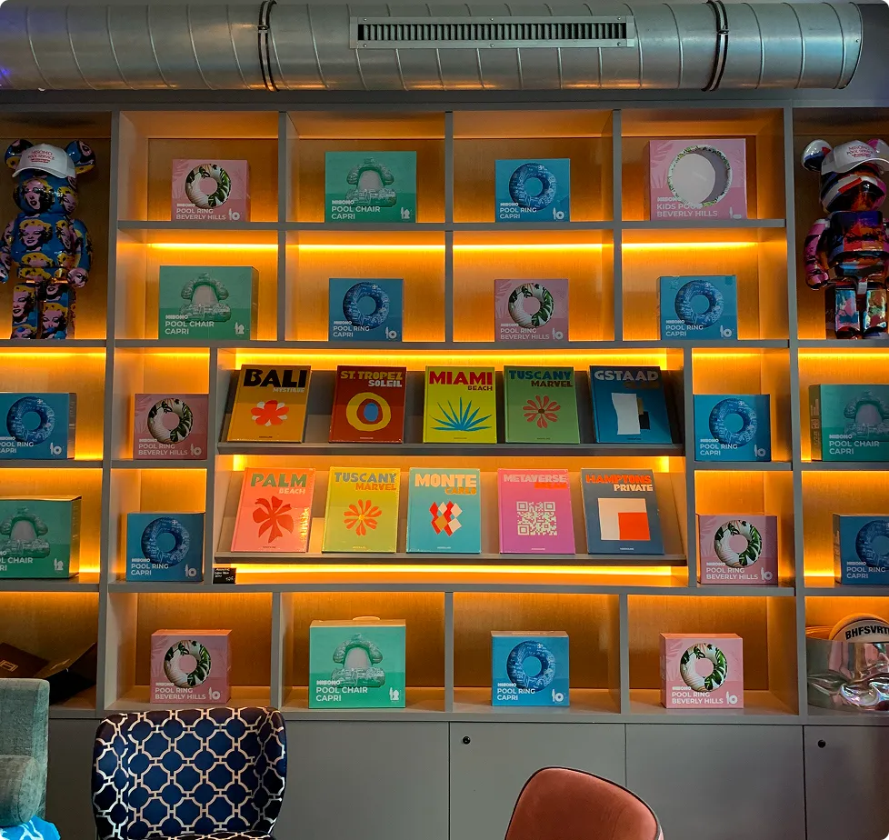 Beleuchtetes Regal mit bunten Büchern zu Reisezielen und Pool-Spielzeugen, flankiert von zwei bunt gemusterten Bärenfiguren mit weißen Kappen.